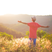 Person wearing a hat and raising arms while standing on a hill overlooking a mountain range