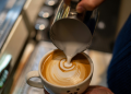 Hand holding container of milk being poured into latte mug