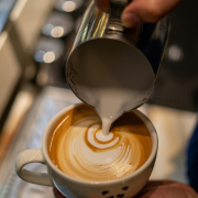 Hand holding container of milk being poured into latte mug