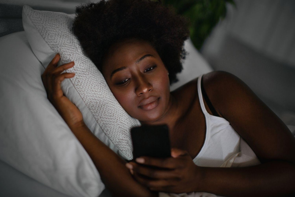 A woman with dark curly hair lies in bed in a dimly lit room, resting her head on a white pillow. She has a thoughtful or neutral expression as she looks down at the glowing screen of a smartphone held in her hand. She is wearing a white tank top, and the scene captures a quiet, late-night atmosphere.