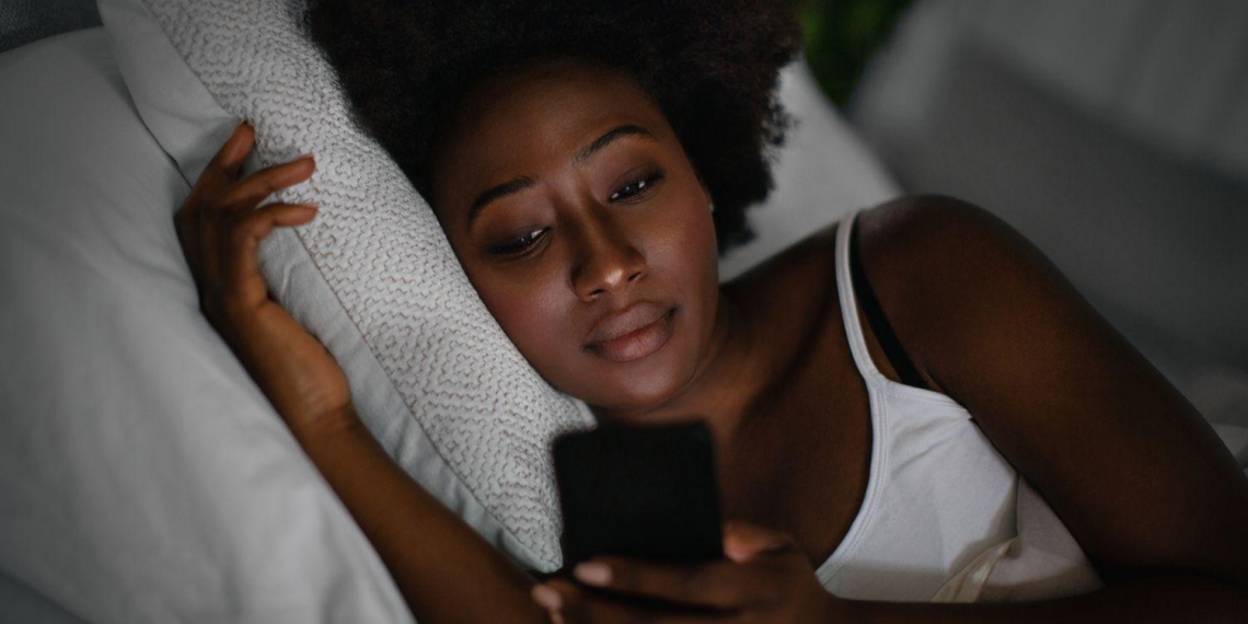 A woman with dark curly hair lies in bed in a dimly lit room, resting her head on a white pillow. She has a thoughtful or neutral expression as she looks down at the glowing screen of a smartphone held in her hand. She is wearing a white tank top, and the scene captures a quiet, late-night atmosphere.