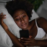 A woman with dark curly hair lies in bed in a dimly lit room, resting her head on a white pillow. She has a thoughtful or neutral expression as she looks down at the glowing screen of a smartphone held in her hand. She is wearing a white tank top, and the scene captures a quiet, late-night atmosphere.