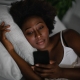 A woman with dark curly hair lies in bed in a dimly lit room, resting her head on a white pillow. She has a thoughtful or neutral expression as she looks down at the glowing screen of a smartphone held in her hand. She is wearing a white tank top, and the scene captures a quiet, late-night atmosphere.