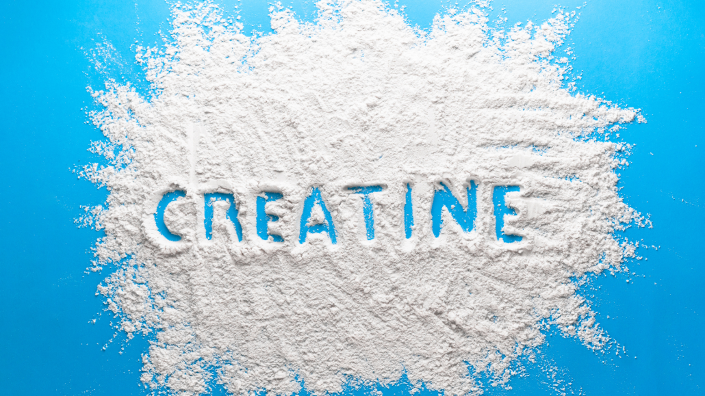 A top-down view of a bright blue surface covered with a scattered pile of fine white powder. The word "CREATINE" is traced into the center of the powder, revealing the blue background underneath. The powder has a textured, grainy appearance with soft edges spreading toward the frame.