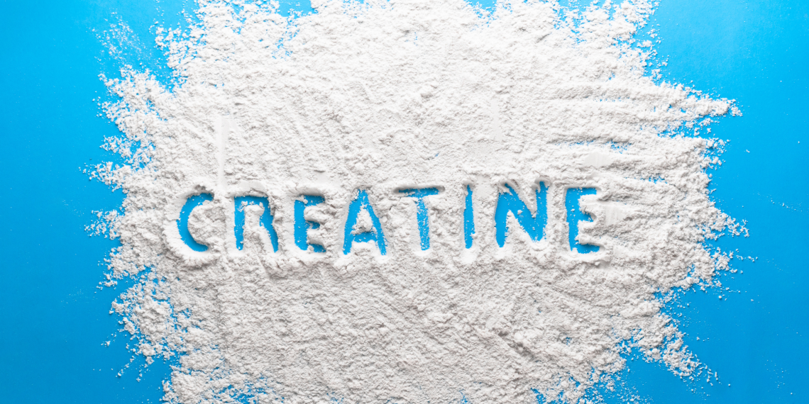 A top-down view of a bright blue surface covered with a scattered pile of fine white powder. The word "CREATINE" is traced into the center of the powder, revealing the blue background underneath. The powder has a textured, grainy appearance with soft edges spreading toward the frame.