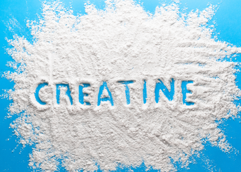 A top-down view of a bright blue surface covered with a scattered pile of fine white powder. The word "CREATINE" is traced into the center of the powder, revealing the blue background underneath. The powder has a textured, grainy appearance with soft edges spreading toward the frame.