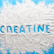 A top-down view of a bright blue surface covered with a scattered pile of fine white powder. The word "CREATINE" is traced into the center of the powder, revealing the blue background underneath. The powder has a textured, grainy appearance with soft edges spreading toward the frame.