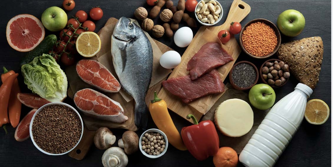 A top-down overhead view of a diverse assortment of fresh, healthy foods arranged on a dark wooden surface. The collection includes proteins like a whole fish, salmon fillets, raw red meat, and eggs; fresh produce such as green apples, grapefruit halves, tomatoes, peppers, and leafy greens; and pantry staples including lentils, chickpeas, nuts, seeds, and a bottle of milk. The vibrant colors of the fruits and vegetables contrast against the dark background, emphasizing a balanced and nutritious diet.