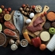 A top-down overhead view of a diverse assortment of fresh, healthy foods arranged on a dark wooden surface. The collection includes proteins like a whole fish, salmon fillets, raw red meat, and eggs; fresh produce such as green apples, grapefruit halves, tomatoes, peppers, and leafy greens; and pantry staples including lentils, chickpeas, nuts, seeds, and a bottle of milk. The vibrant colors of the fruits and vegetables contrast against the dark background, emphasizing a balanced and nutritious diet.