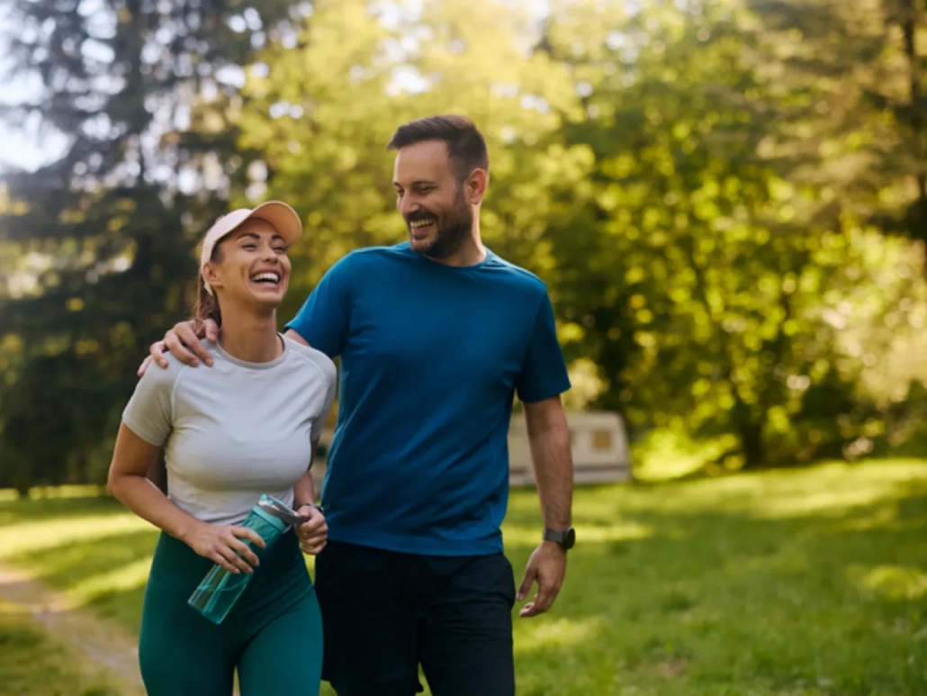 A man and a woman are walking together through a sun-drenched park, both smiling and laughing. The man, wearing a blue t-shirt and black shorts, has his arm around the woman's shoulder. She is wearing a light grey t-shirt, dark teal leggings, and a peach-colored visor, and is holding a translucent blue water bottle. The lush green trees in the background are bathed in warm, soft sunlight, creating a bright and energetic atmosphere.