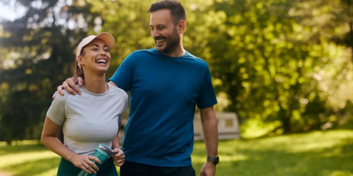 A man and a woman are walking together through a sun-drenched park, both smiling and laughing. The man, wearing a blue t-shirt and black shorts, has his arm around the woman's shoulder. She is wearing a light grey t-shirt, dark teal leggings, and a peach-colored visor, and is holding a translucent blue water bottle. The lush green trees in the background are bathed in warm, soft sunlight, creating a bright and energetic atmosphere.