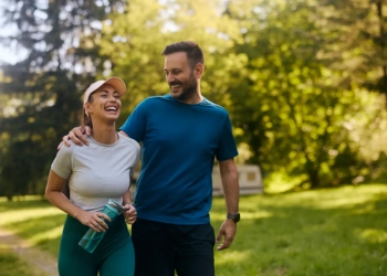 A man and a woman are walking together through a sun-drenched park, both smiling and laughing. The man, wearing a blue t-shirt and black shorts, has his arm around the woman's shoulder. She is wearing a light grey t-shirt, dark teal leggings, and a peach-colored visor, and is holding a translucent blue water bottle. The lush green trees in the background are bathed in warm, soft sunlight, creating a bright and energetic atmosphere.