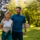 A man and a woman are walking together through a sun-drenched park, both smiling and laughing. The man, wearing a blue t-shirt and black shorts, has his arm around the woman's shoulder. She is wearing a light grey t-shirt, dark teal leggings, and a peach-colored visor, and is holding a translucent blue water bottle. The lush green trees in the background are bathed in warm, soft sunlight, creating a bright and energetic atmosphere.