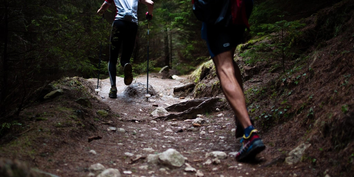 A low-angle, action shot from behind shows two trail runners navigating a rugged, rocky path through a dense, dark forest. The runner in the lead is using trekking poles and wearing a white and red jersey with black leggings, while the runner in the foreground is captured mid-stride, showcasing muscular legs and colorful trail shoes. The earthy terrain is covered in stones and roots, surrounded by deep green evergreen trees that create a moody, adventurous atmosphere.
