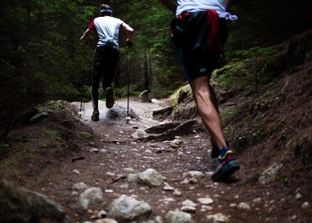 A low-angle, action shot from behind shows two trail runners navigating a rugged, rocky path through a dense, dark forest. The runner in the lead is using trekking poles and wearing a white and red jersey with black leggings, while the runner in the foreground is captured mid-stride, showcasing muscular legs and colorful trail shoes. The earthy terrain is covered in stones and roots, surrounded by deep green evergreen trees that create a moody, adventurous atmosphere.