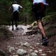A low-angle, action shot from behind shows two trail runners navigating a rugged, rocky path through a dense, dark forest. The runner in the lead is using trekking poles and wearing a white and red jersey with black leggings, while the runner in the foreground is captured mid-stride, showcasing muscular legs and colorful trail shoes. The earthy terrain is covered in stones and roots, surrounded by deep green evergreen trees that create a moody, adventurous atmosphere.