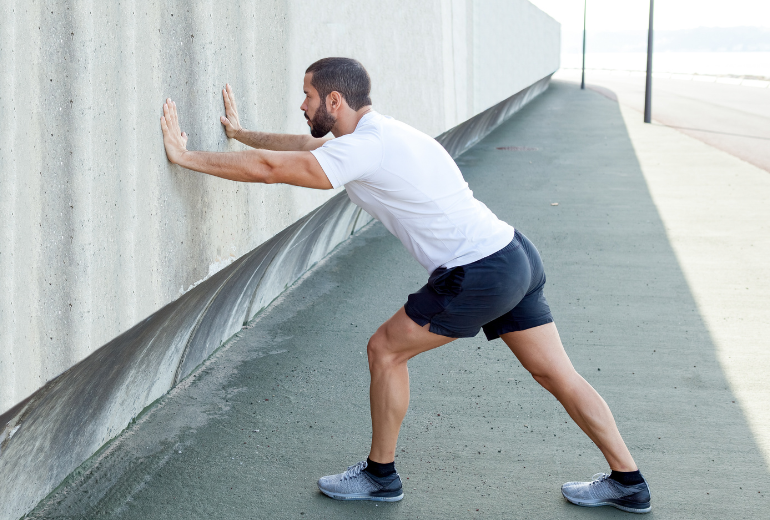 Man stretching his calves by pressing arms against wall of building