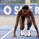 A female track athlete with a focused expression is positioned in starting blocks on a blue running track. She is wearing a red athletic top and white racing shoes, with her hands planted firmly on the track surface. In the blurred background, a blue banner with large white lettering is visible behind a fence.