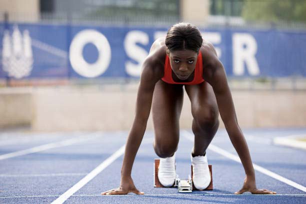 A female track athlete with a focused expression is positioned in starting blocks on a blue running track. She is wearing a red athletic top and white racing shoes, with her hands planted firmly on the track surface. In the blurred background, a blue banner with large white lettering is visible behind a fence.