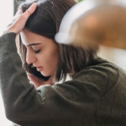 A young woman with a short brown bob hairstyle appears stressed or overwhelmed while talking on a smartphone. She is wearing a textured olive green sweater and has one hand pressed against her forehead as she looks downward. The image is a close-up profile shot with a soft, warm-toned lamp blurred in the foreground, creating a shallow depth of field.