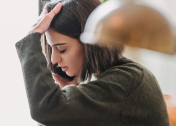 A young woman with a short brown bob hairstyle appears stressed or overwhelmed while talking on a smartphone. She is wearing a textured olive green sweater and has one hand pressed against her forehead as she looks downward. The image is a close-up profile shot with a soft, warm-toned lamp blurred in the foreground, creating a shallow depth of field.