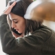 A young woman with a short brown bob hairstyle appears stressed or overwhelmed while talking on a smartphone. She is wearing a textured olive green sweater and has one hand pressed against her forehead as she looks downward. The image is a close-up profile shot with a soft, warm-toned lamp blurred in the foreground, creating a shallow depth of field.