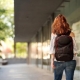 A woman with long, wavy reddish-brown hair is shown from the back, walking down an outdoor corridor or walkway alongside a modern building with large glass windows. She is wearing an off-the-shoulder striped top, denim jeans, and a black backpack with red accents. To her left, there is a row of lush green trees, and the background has a soft, blurred depth of field.