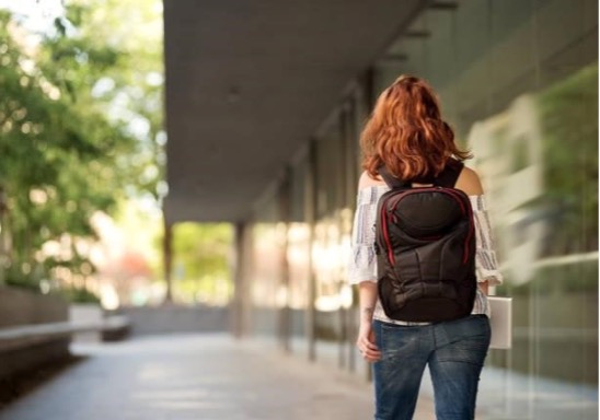 A woman with long, wavy reddish-brown hair is shown from the back, walking down an outdoor corridor or walkway alongside a modern building with large glass windows. She is wearing an off-the-shoulder striped top, denim jeans, and a black backpack with red accents. To her left, there is a row of lush green trees, and the background has a soft, blurred depth of field.