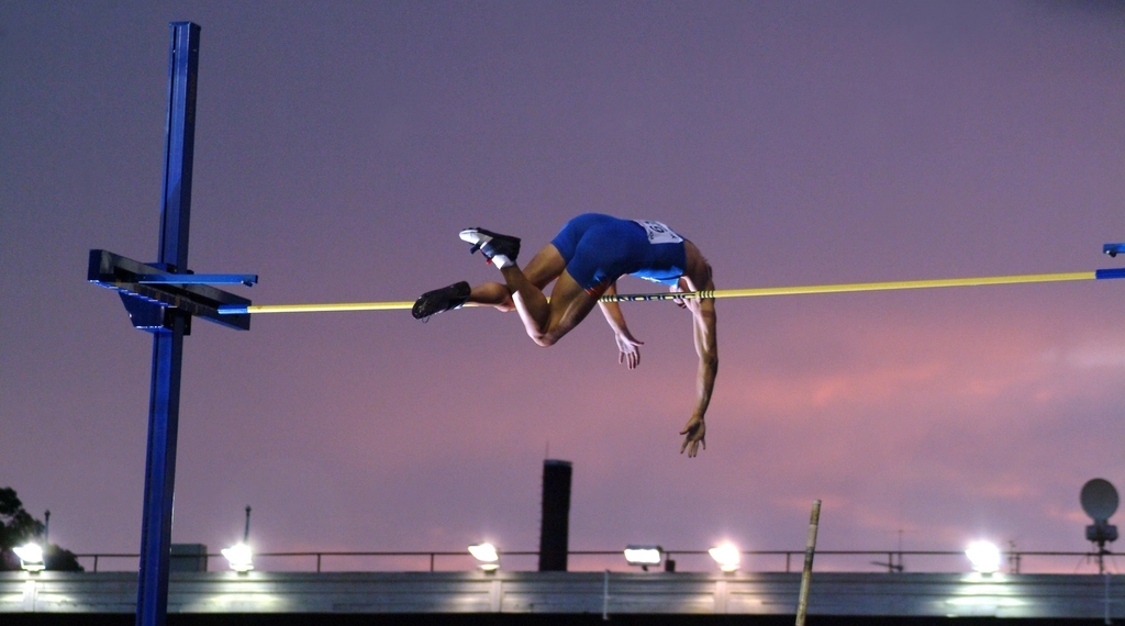 A pole vaulter in a blue singlet is captured in mid-air, arching their body over a yellow horizontal bar against a twilight sky. The background shows a stadium structure with bright lights and a purple and orange sunset glow.