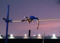 A pole vaulter in a blue singlet is captured in mid-air, arching their body over a yellow horizontal bar against a twilight sky. The background shows a stadium structure with bright lights and a purple and orange sunset glow.