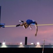 A pole vaulter in a blue singlet is captured in mid-air, arching their body over a yellow horizontal bar against a twilight sky. The background shows a stadium structure with bright lights and a purple and orange sunset glow.