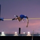 A pole vaulter in a blue singlet is captured in mid-air, arching their body over a yellow horizontal bar against a twilight sky. The background shows a stadium structure with bright lights and a purple and orange sunset glow.