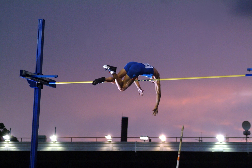 A pole vaulter in a blue singlet is captured in mid-air, arching their body over a yellow horizontal bar against a twilight sky. The background shows a stadium structure with bright lights and a purple and orange sunset glow.