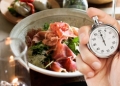 A person's hand holds a silver mechanical stopwatch in the foreground, with the thumb ready to press the top button. In the blurred background, a healthy meal is visible, featuring a bowl filled with leafy greens and slices of prosciutto. The composition suggests the concept of timed eating or intermittent fasting, contrasting the precision of the clock with the act of dining.