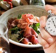 A person's hand holds a silver mechanical stopwatch in the foreground, with the thumb ready to press the top button. In the blurred background, a healthy meal is visible, featuring a bowl filled with leafy greens and slices of prosciutto. The composition suggests the concept of timed eating or intermittent fasting, contrasting the precision of the clock with the act of dining.