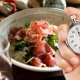 A person's hand holds a silver mechanical stopwatch in the foreground, with the thumb ready to press the top button. In the blurred background, a healthy meal is visible, featuring a bowl filled with leafy greens and slices of prosciutto. The composition suggests the concept of timed eating or intermittent fasting, contrasting the precision of the clock with the act of dining.