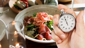 A person's hand holds a silver mechanical stopwatch in the foreground, with the thumb ready to press the top button. In the blurred background, a healthy meal is visible, featuring a bowl filled with leafy greens and slices of prosciutto. The composition suggests the concept of timed eating or intermittent fasting, contrasting the precision of the clock with the act of dining.