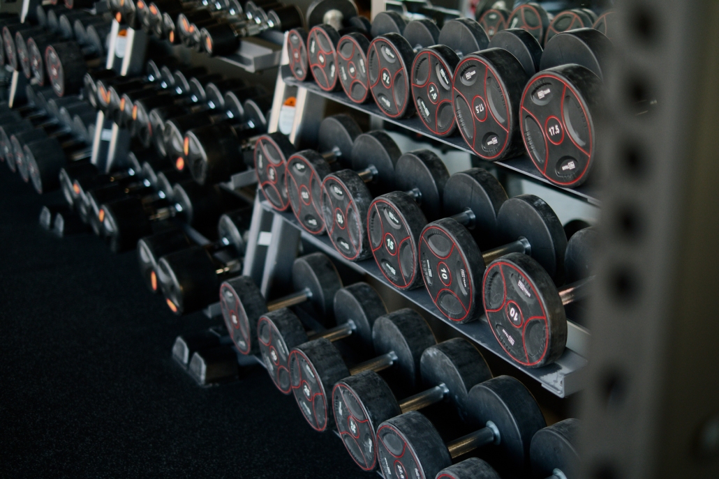 A perspective shot of a gym weight rack filled with multiple rows of black dumbbells. The weights feature red circular accents and numerical labels indicating their weight in kilograms. The rack recedes into the background, creating a sense of depth and variety, highlighting the range of equipment available for strength training.