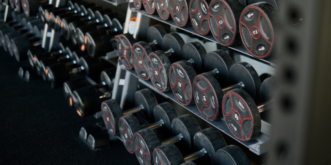 A perspective shot of a gym weight rack filled with multiple rows of black dumbbells. The weights feature red circular accents and numerical labels indicating their weight in kilograms. The rack recedes into the background, creating a sense of depth and variety, highlighting the range of equipment available for strength training.