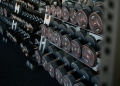 A perspective shot of a gym weight rack filled with multiple rows of black dumbbells. The weights feature red circular accents and numerical labels indicating their weight in kilograms. The rack recedes into the background, creating a sense of depth and variety, highlighting the range of equipment available for strength training.