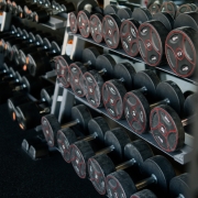 A perspective shot of a gym weight rack filled with multiple rows of black dumbbells. The weights feature red circular accents and numerical labels indicating their weight in kilograms. The rack recedes into the background, creating a sense of depth and variety, highlighting the range of equipment available for strength training.