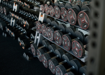 A perspective shot of a gym weight rack filled with multiple rows of black dumbbells. The weights feature red circular accents and numerical labels indicating their weight in kilograms. The rack recedes into the background, creating a sense of depth and variety, highlighting the range of equipment available for strength training.